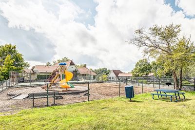 Fenced in playground with slide next to a picnic area with bench trash can and picnic table on a grass area.
